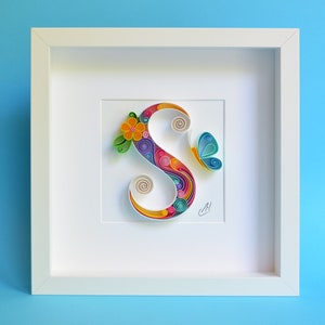 May include: A white frame with a colorful paper art letter S, a butterfly, and a flower. The letter S is made of curled paper strips in shades of pink, blue, green, yellow, and orange. The butterfly is blue and white, and the flower is yellow with green leaves.