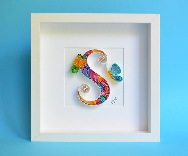 May include: A white frame with a colorful paper art letter S, a butterfly, and a flower. The letter S is made of curled paper strips in shades of pink, blue, green, yellow, and orange. The butterfly is blue and white, and the flower is yellow with green leaves.