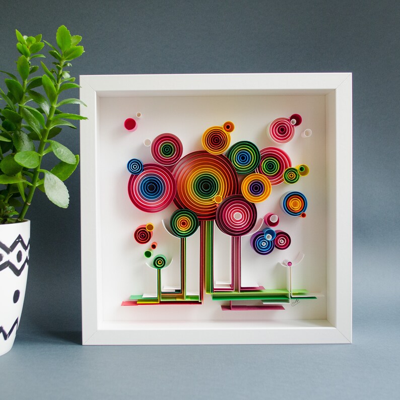 May include: A framed paper art piece featuring a colorful, abstract design. The artwork showcases concentric circles in various sizes and rainbow hues, resembling stylized trees. The white frame provides a clean border, enhancing the vibrant colors of the artwork.