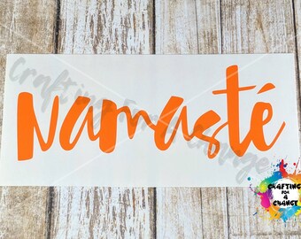 May include: Orange vinyl decal with the word "Namaste" in a cursive font. The decal is on a white background, with a colorful paint splatter design in the bottom right corner. The background is a light wood grain.
