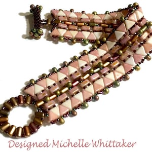 Op de afbeelding: Een roze, witte en bruine kralen armband met een geometrisch patroon. De armband heeft een sluiting en een ring aan het einde. "Designed Michelle Whittaker" staat onder de armband.