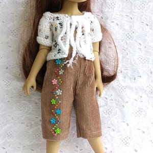 Puede incluir: Una muñeca vestida con una rebeca de punto blanca con detalles florales, pantalones capri de pana marrón adornados con bordados florales coloridos y zapatos Mary Jane rosas. La muñeca tiene el pelo largo castaño y está sobre un fondo blanco.