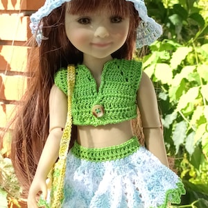 Peut inclure: Une poupée vêtue d'un ensemble crocheté fait à la main. La tenue comprend un haut vert avec un bouton, une jupe bleue et blanche et un chapeau assorti. Un petit sac jaune est suspendu à l'épaule de la poupée. La poupée a de longs cheveux bruns.