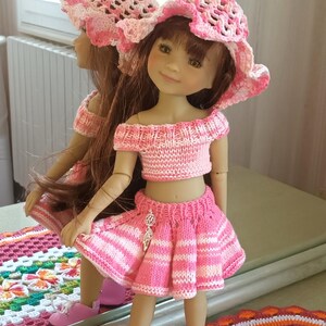Peut inclure: Une poupée aux longs cheveux bruns portant un haut en tricot à rayures roses et blanches, une jupe plissée assortie avec une breloque argentée et un chapeau de soleil crocheté rose. La poupée porte des chaussures roses à lacets blancs.