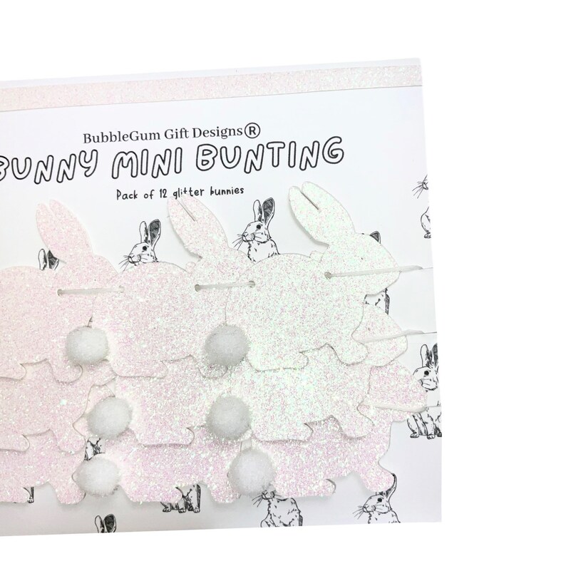 K&ouml;nnte beinhalten: Eine Packung mit 12 wei&szlig;en Glitzer-Hasen-Mini-Girlanden. Die Hasen sind mit wei&szlig;em Garn aneinandergereiht und haben wei&szlig;e Bommel dazwischen. Die Verpackung lautet "BubbleGum Gift Designs&reg; Bunny Mini Bunting Pack of 12 glitter bunnies".
