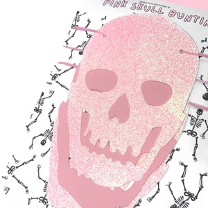 May include: Pink glitter skull bunting with a white background featuring a repeating pattern of black skeletons. The bunting is made of pink glitter paper and has a white string running through it.