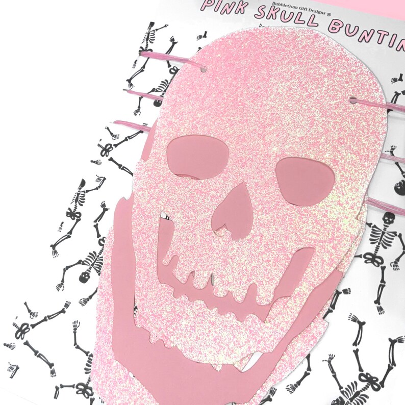 May include: Pink glitter skull bunting with a white background featuring a repeating pattern of black skeletons. The bunting is made of pink glitter paper and has a white string running through it.