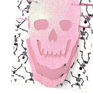 May include: Pink glitter skull Halloween banner with a layered design. The banner is decorated with a repeating pattern of black skeletons.