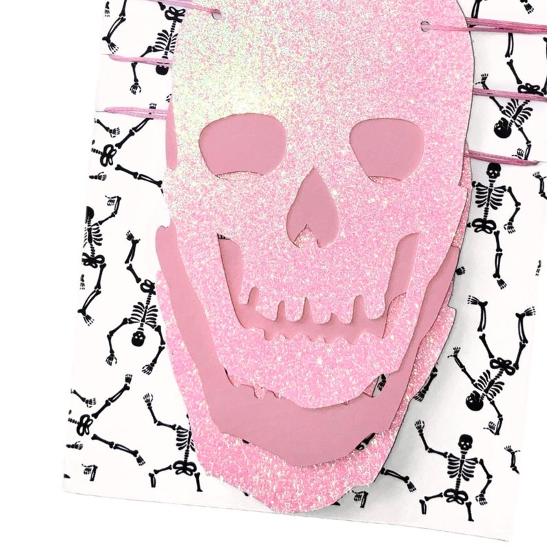 May include: Pink glitter skull Halloween banner with a layered design. The banner is decorated with a repeating pattern of black skeletons.