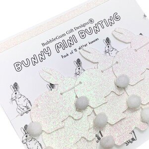 K&ouml;nnte beinhalten: Wei&szlig;er Glitzer-Hasen-Mini-Girlande mit 12 Hasen und wei&szlig;en Pompons. Die Verpackung lautet "BubbleGum Gift Designs&reg; Bunny Mini Bunting Pack of 12 glitter bunnies".