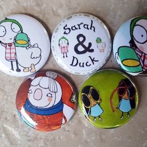 May include: Five round buttons with colorful cartoon illustrations. The buttons feature a girl with a duck, a girl with sunglasses, a girl with a red scarf, and two buttons with the text "Sarah & Duck".