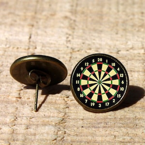 Puede incluir: Un par de pendientes de botón de color bronce con un diseño de diana de dardos. La diana de dardos es negra y blanca con detalles rojos y verdes. Los números del 1 al 20 están impresos alrededor de la diana de dardos.