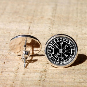 Puede incluir: Un par de pendientes de botón de plata con fondo negro y un diseño de brújula blanco rodeado de runas.