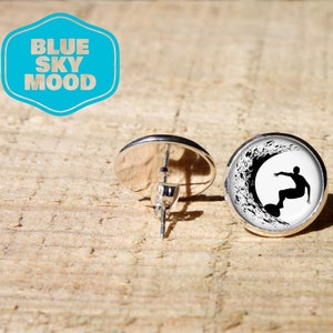 Puede incluir: Pendientes de botón plateados con un diseño de surfista en blanco y negro. Los pendientes tienen un diseño circular y un acabado brillante. La imagen también incluye un logotipo azul claro y blanco que dice "BLUE SKY MOOD".