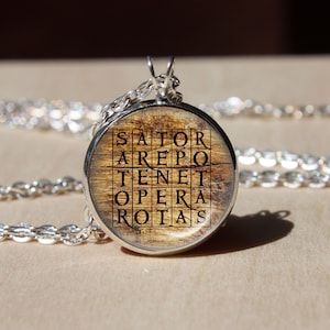Puede incluir: Un collar de cadena de plata con un colgante redondo con un fondo de madera y el cuadrado de palabras latino "SATOR AREPO TENET OPERA ROTAS" impreso en negro.