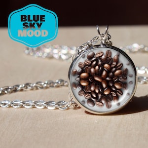Op de afbeelding: Een zilverkleurige ketting met een ronde hanger gevuld met koffiebonen. De hanger hangt aan een fijne ketting. Een blauw-wit logo met de tekst "BLUE SKY MOOD" staat linksboven.