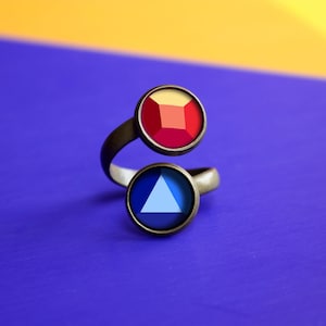 May include: A bronze-toned ring with two circular settings. The top setting features a red and orange square gemstone, while the bottom setting has a blue triangle gemstone. The ring has a unique, open design.