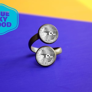 Może przedstawiać: Srebrny, regulowany pierścionek z dwoma okrągłymi, lustrzanymi zawieszkami w stylu dyskotekowej kuli. Pierścionek leży na niebieskiej powierzchni z żółtą powierzchnią w tle. Tekst "BLUE SKY MOOD" znajduje się w niebieskim kółku w lewym górnym rogu obrazu.