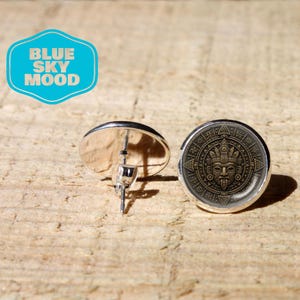 May include: A pair of round stud earrings featuring a detailed bronze-toned Aztec calendar design on the front. The earrings have a silver-toned metal post backing. A blue logo graphic with the text BLUE SKY MOOD is visible in the upper left corner.