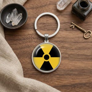 Könnte beinhalten: Ein runder Schlüsselanhänger mit einem gelb-schwarzen Strahlungsgefahrensymbol, eingefasst in einen silberfarbenen Metallrahmen mit passendem Schlüsselring. Das Design zeigt das ikonische Warnzeichen mit drei schwarzen Flügeln.
