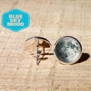 May include: A pair of round stud earrings featuring a detailed photographic print of the moon's cratered surface in shades of grey and white. The earrings have a silver-toned metal setting and a post back. A blue logo reads BLUE SKY MOOD.