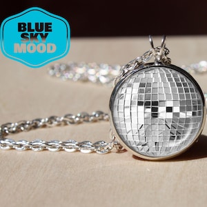 Peut inclure: Collier argenté avec un pendentif rond en forme de boule à facettes, composé de petits carrés miroités. La chaîne est fine. Le logo bleu et blanc "BLUE SKY MOOD" est également visible.