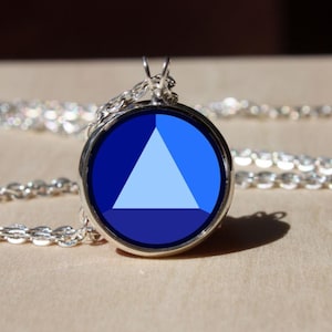 Könnte beinhalten: Halskette mit silberfarbenem Anhänger, der ein kreisförmiges blaues Design mit einem hellblauen Dreieck in der Mitte aufweist. Der Anhänger hängt an einer filigranen Kette und bildet ein schlichtes, aber auffälliges Schmuckstück.