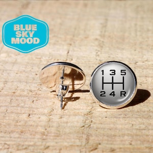 May include: A pair of round stud earrings featuring a black manual transmission gear shift pattern on a white background. One earring shows the front face with the numbers 1, 3, 5, 2, 4, and R, while the other shows the silver metal post backing.
