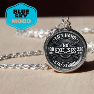 May include: A silver necklace with a round pendant featuring a weightlifting design. The pendant has the text "LIFT HARD, NO EXCUSES, STAY STRONG" and measurements of 100 KG and 220 LBS. A blue and white logo is in the upper left corner.