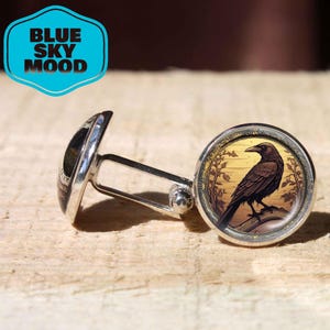 Puede incluir: Gemelos plateados con una ilustración detallada de un cuervo negro posado en una rama, sobre un fondo dorado. Los gemelos tienen una cara redonda y una barra plateada. El texto "BLUE SKY MOOD" está en un hexágono azul.