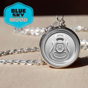 Op de afbeelding: Een zilverkleurige hangerketting met een miniatuur frisdrankblikje. De hanger is bevestigd aan een fijne ketting. De afbeelding bevat ook een lichtblauw en wit logo met de tekst "BLUE SKY MOOD".