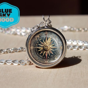 Puede incluir: Colgante de brújula plateado con una esfera detallada en oro y azul, suspendido de una cadena plateada. La imagen incluye el texto "BLUE SKY HOOD" en un diseño azul y blanco. El diseño de la brújula evoca un tema náutico.