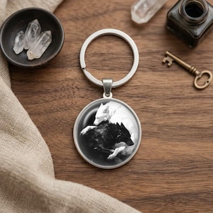 May include: A circular metal keychain featuring a black and white graphic illustration of two wolves, one white and one black, positioned in a yin and yang style composition. The keychain has a silver-toned metal ring and a circular pendant frame.