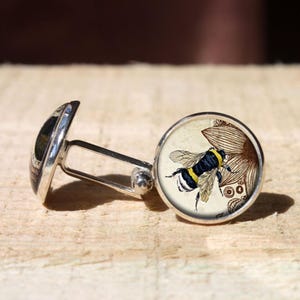 May include: Silver cufflinks with a round face featuring a detailed illustration of a bumblebee with yellow and black stripes near a brown flower. The cufflinks are on a light brown wooden surface.