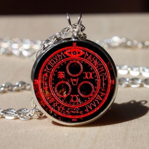 Pode incluir: Colar com pingente prateado. O pingente circular tem um fundo preto com um design vermelho detalhado com símbolos e texto. A corrente também é prateada.