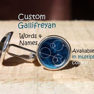 Puede incluir: Gemelos plateados con una cara redonda azul con un diseño geométrico blanco. El texto "Custom Gallifreyan Words & Names" es visible. Disponibles en varios colores.