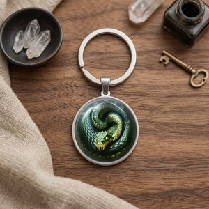 Peut inclure: Un porte-clés circulaire en métal doté d'un cabochon en verre présentant une illustration détaillée d'un serpent vert enroulé. Le serpent possède des écailles texturées, des yeux jaunes et des crocs visibles sur un fond sombre et ombragé.