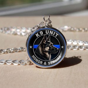 May include: A silver-toned pendant necklace featuring a circular black emblem with a German Shepherd profile. The design includes a horizontal blue line, two white stars, and the text K9 UNIT HONOR & SERVISE in white capital letters.