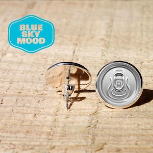 Puede incluir: Un par de pendientes de botón plateados. Uno presenta una superficie reflectante, mientras que el otro está diseñado para parecerse a la parte superior de una lata de refresco. Los pendientes están sobre un fondo de madera. Un logotipo azul y blanco que dice "BLUE SKY MOOD" está en el fondo.