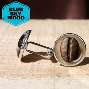 May include: Silver cufflinks with a coffee bean design. One cufflink features a detailed coffee bean, while the other has a dark, patterned design. The cufflinks are connected by a silver bar and a small ball.