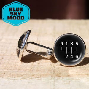 Könnte beinhalten: Silberne Manschettenknöpfe mit schwarzem Zifferblatt und weißem Schaltschema. Das Schaltschema umfasst R, 1, 3, 5, 2, 4 und 6. Die Manschettenknöpfe liegen auf einer hellbraunen Holzoberfläche. Der Text "BLUE SKY MOOD" befindet sich in einem blauen Badge.