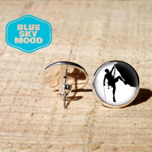 Peut inclure: Boucles d'oreilles à clous rondes avec une monture en métal argenté. Une boucle présente une silhouette noire d'une personne faisant de l'escalade sur fond blanc. L'autre boucle est vue de dos, révélant la tige et le fermoir papillon.