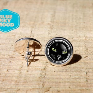 Peut inclure: Boucles d'oreilles à clous argentées avec un motif de visage de chat noir. Le chat a des yeux verts et des moustaches blanches. Les boucles d'oreilles sont rondes et reposent sur une surface en bois brun clair. Le texte "BLUE SKY MOOD" est dans un hexagone bleu.