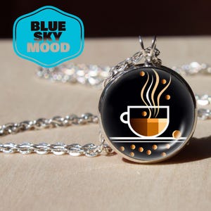 Puede incluir: Un collar plateado con un colgante redondo con un diseño de taza de café. La taza es marrón y blanca, con vapor saliendo. El colgante está sobre un fondo negro. El texto "BLUE SKY MOOD" está en un hexágono azul y blanco.