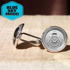 Puede incluir: Gemelos plateados con diseño de lata. Un gemelo presenta la parte superior de una lata, el otro tiene un diseño negro y plateado. Los gemelos están sobre una superficie de madera. El texto "BLUE SKY MOOD" está en una insignia azul y blanca.