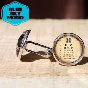 May include: Silver cufflinks with a round face featuring an eye chart design. The eye chart has the letter "H" at the top and the words "BLUE SKY MOOD" in a blue and white logo.