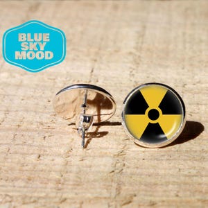Peut inclure: Une paire de boucles d'oreilles à clous argentées. L'une des boucles d'oreilles affiche un symbole de radiation noir et jaune. L'autre boucle d'oreille est tournée sur le côté, montrant la tige et le dos. Le fond est une surface en bois brun clair. Un logo bleu et blanc avec "BLUE SKY MOOD" est dans le coin supérieur gauche.