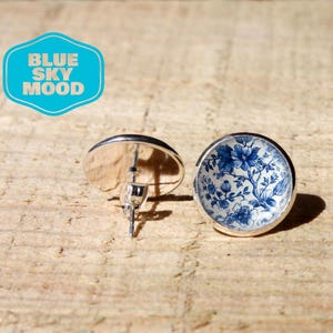 May include: A pair of round stud earrings with a floral design in shades of blue and white. The earrings have a silver-colored metal setting. The image includes a blue and white logo that says "BLUE SKY MOOD".
