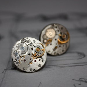 May include: Two silver-toned cufflinks with intricate watch mechanism details. The gears, springs, and jewels are visible, with gold and red accents. The cufflinks are round and sit on a gray surface with faint sketches.
