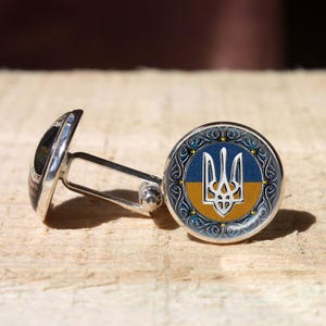 Könnte beinhalten: Silberne Manschettenknöpfe mit einem runden Gesicht, das das ukrainische Wappen in Blau und Gelb zeigt. Das Design ist von einem dekorativen Rand umgeben. Die Manschettenknöpfe liegen auf einer hellbraunen, strukturierten Oberfläche.